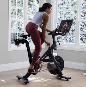 A woman on an indoor exercise bike, cycling while standing on the pedals, her hand turning a dial to adjust the difficulty.